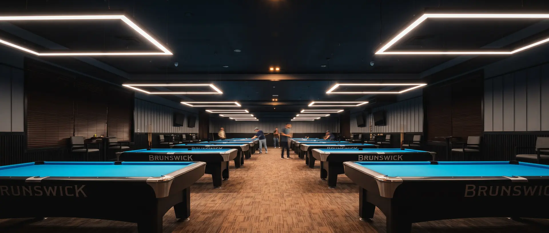 The game floor at Komic Karma — rows of Brunswick tables with electric blue felt under angular LED ceiling fixtures