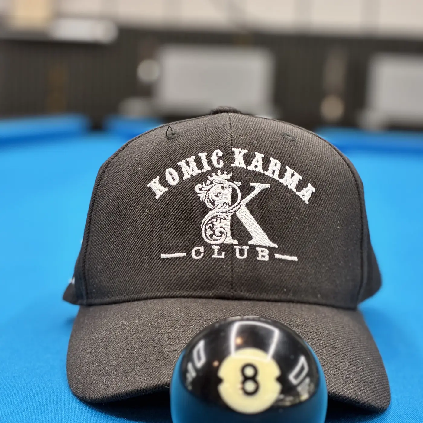 A Komic Karma Club cap resting on the blue felt beside the eight ball